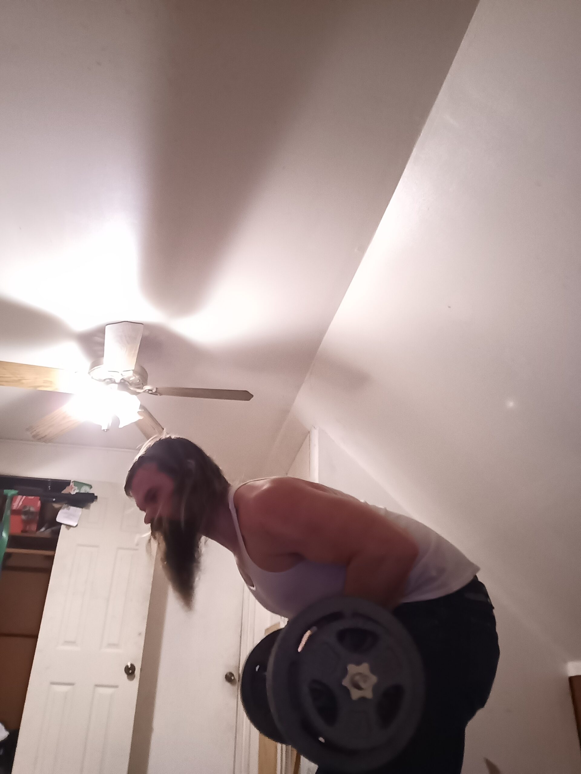 Person in a white tank top lifting a weight plate in a home gym, with a ceiling fan and closet doors visible in the background.