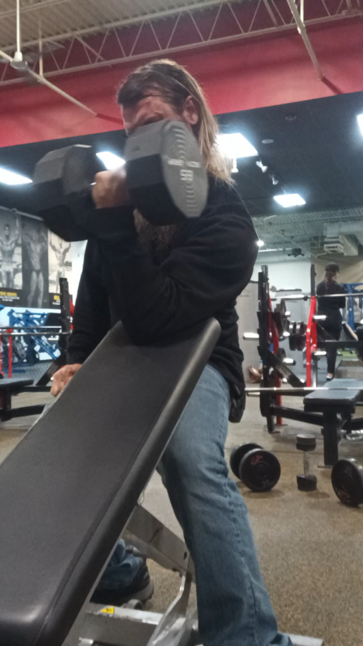 Person in a gym lifting a large dumbbell while seated on an incline bench, wearing a black long-sleeve shirt and jeans.