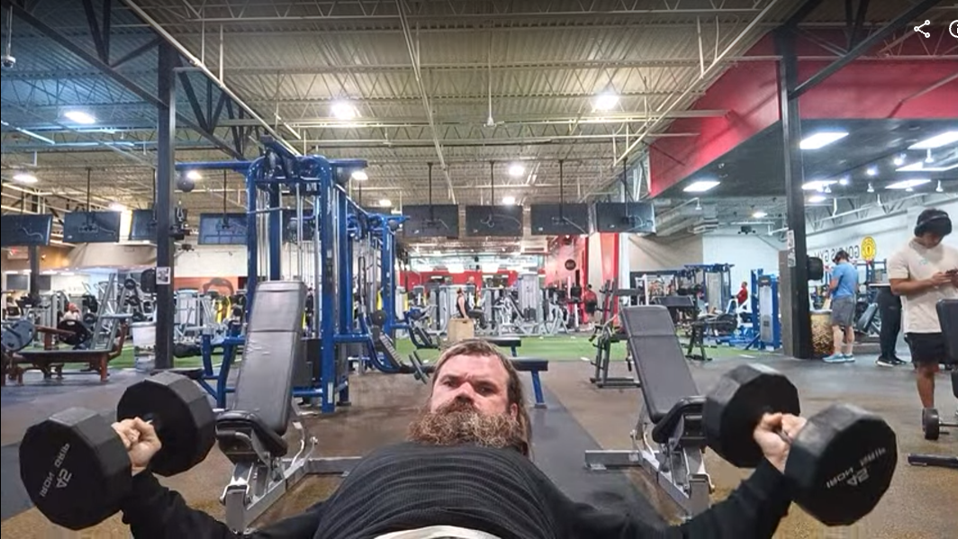 Bearded man bench-pressing two dumbbells on a flat bench in a busy gym, with weight machines in the background.