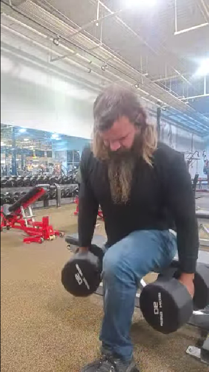 Bearded man in a black shirt lifting two dumbbells in a gym.