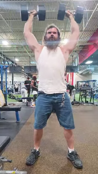 Bearded man in a white tank top and blue shorts lifting two heavy dumbbells overhead in a crowded gym.